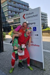 Zwei Clowninnen stehen vor dem Klinikeingang in Lüdenscheid. Eine Clownin trägt die andere Huckepack